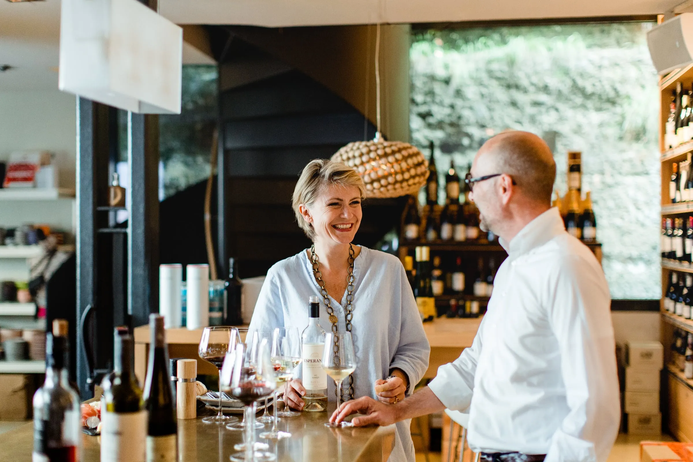 Gastgeberin und Gast im Gespräch an der Bar der Vinothek in der Augustinergasse, auf dem Tresen Weiß- und Rotweingläser, im Hintergrund Weinregale